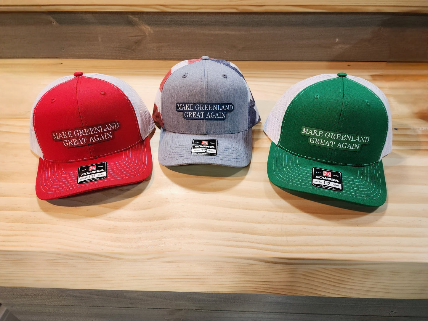 Three "Make Greenland Great Again" hats in red, gray, and green displayed on a wooden surface.