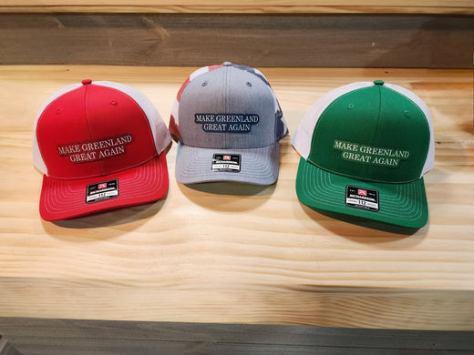 Three "Make Greenland Great Again" hats in red, gray, and green displayed on a wooden surface.