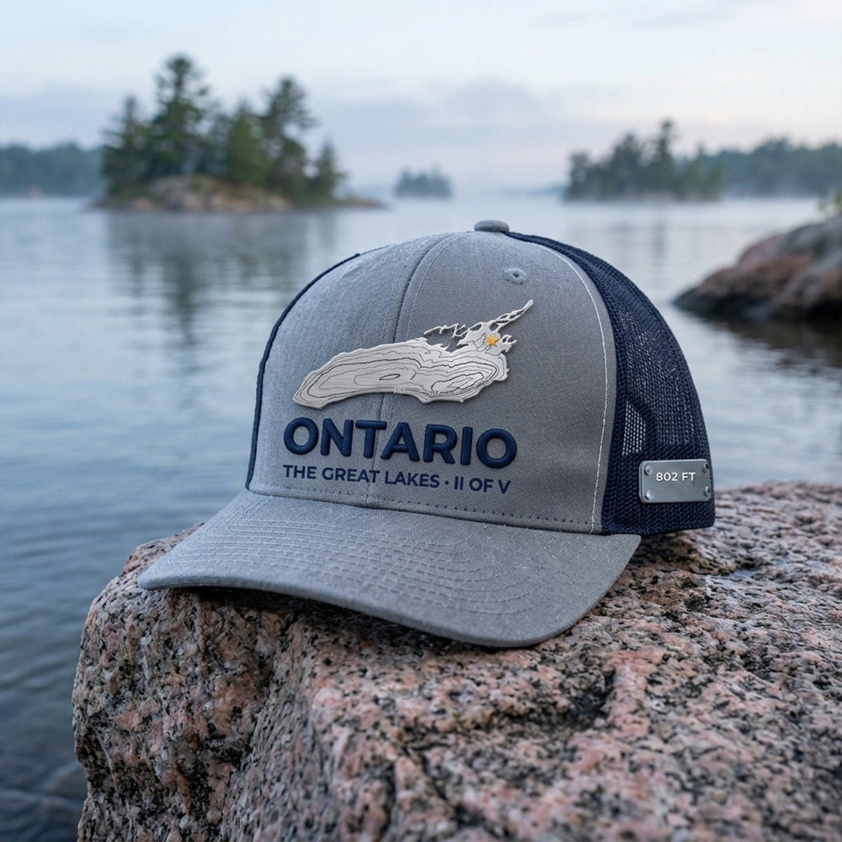 The ONTARIO hat, II of V in The Great Lakes Collector's Edition: a heather grey and navy Richardson 112 trucker cap with a brushed stainless Lake Ontario plate, engraved depth contours and a gold star at the east end, ONTARIO in raised navy lettering over THE GREAT LAKES · II OF V, and an 802 FT metal tag on the side, on a granite rock by the water