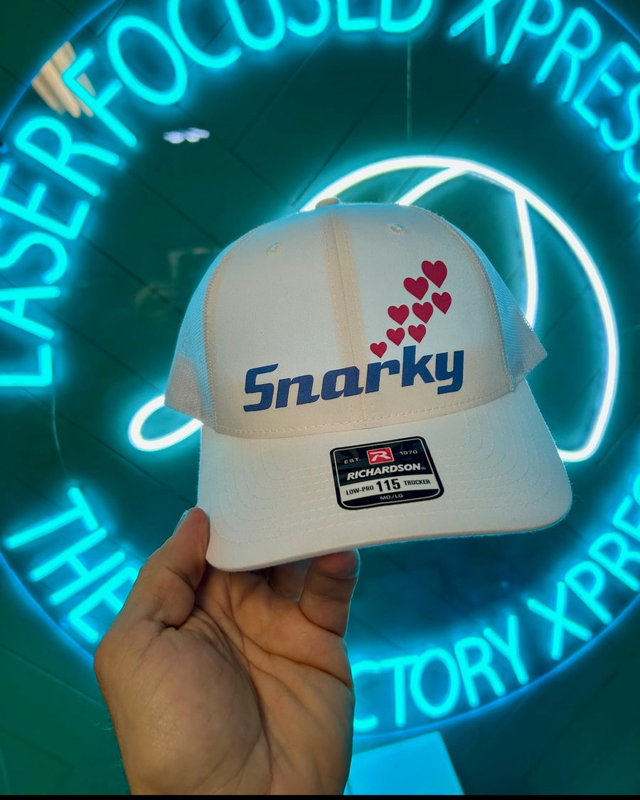 White low-profile trucker hat with a blue script logo and a trail of pink hearts, in front of a neon sign