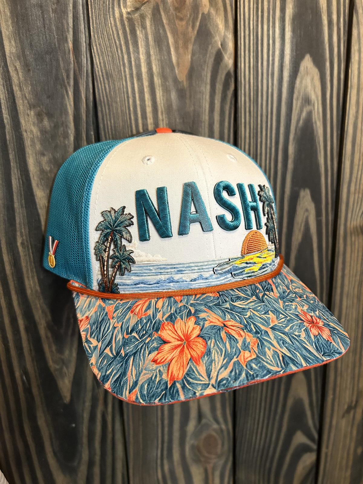 The finished NASH trucker hat on a wood wall: raised teal NASH lettering, palm trees and a sunset ocean scene on a white front, teal mesh sides, an orange rope, a hibiscus-print brim, and a gold medal on the side panel.