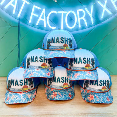 Six identical finished NASH trucker hats stacked under the neon Hat Factory Xpress sign, each with the same teal NASH lettering, palm trees, sunset and hibiscus brim.