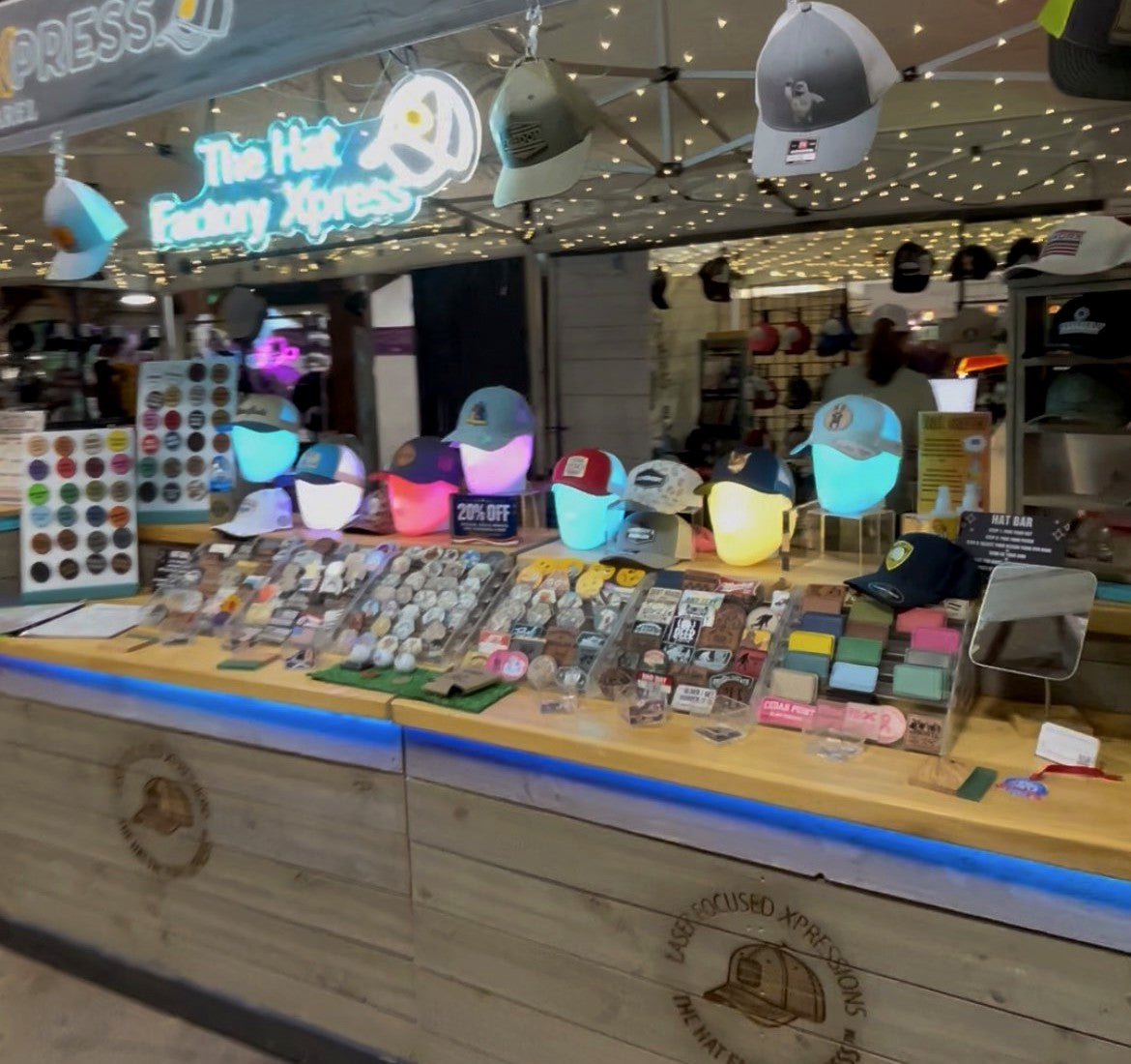 The Hat Factory Xpress pop-up hat bar, set up under its neon sign with caps hanging above the branded wooden counter and trays of patches and ball markers