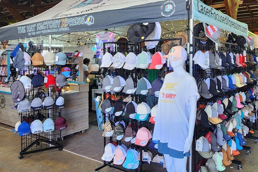 The Hat Factory Xpress® booth at an indoor event: racks of caps in dozens of colours beside the branded canopy and counter