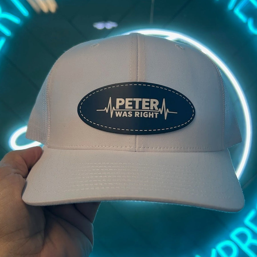 White trucker hat with an engraved navy oval leather patch reading PETER WAS RIGHT with a heartbeat line, held up in front of a neon sign