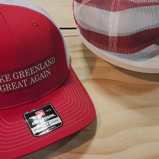 Three "Make Greenland Great Again" hats in red, gray, and green displayed on a wooden surface.