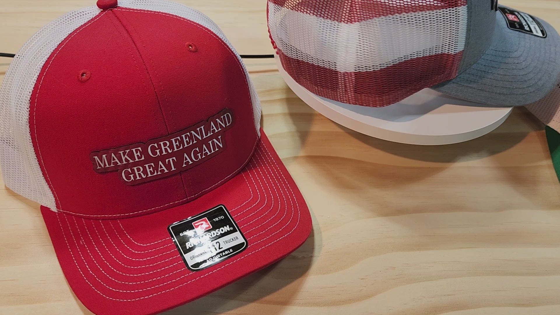 Three "Make Greenland Great Again" hats in red, gray, and green displayed on a wooden surface.