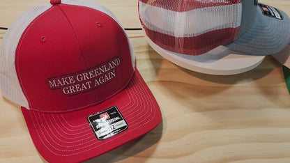 Three "Make Greenland Great Again" hats in red, gray, and green displayed on a wooden surface.